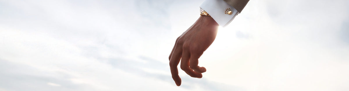Hand with a white shirt cuff wearing Rolex crown cufflinks and a gold Rolex watch against the sky